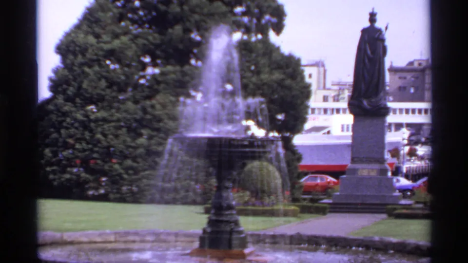 1974 Canada 8mm frame showing a park fountain spraying beside a statue, with trees, lawn, buildings, and cars beyond.