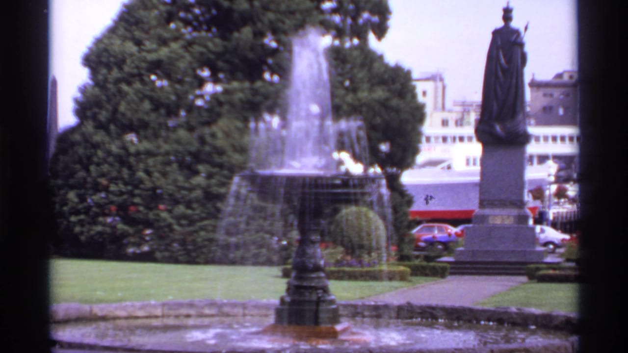 Fountain spraying beside a robed statue in a landscaped urban garden.