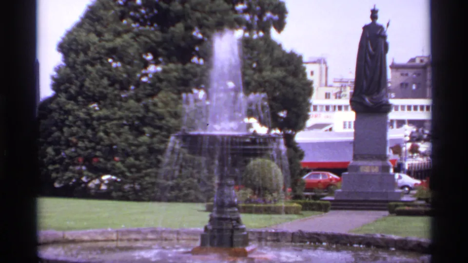 1974 Victoria travel frame with fountain spray, a robed statue on a pedestal, lawns, trees, traffic, and buildings behind.