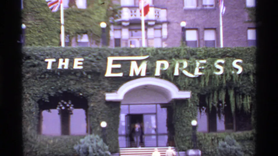 The Empress Hotel entrance in Victoria, British Columbia, with large white lettering, ivy, flags, arched doorway, and people near the doors.