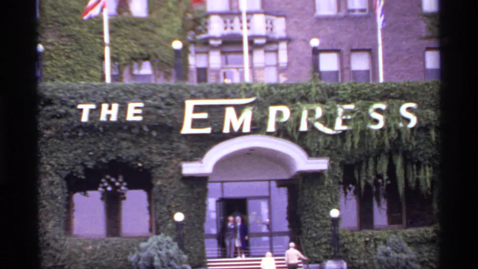 1974 8mm frame of The Empress Hotel exterior in Victoria, with signage, ivy, flags, entrance doors, and visible visitors.