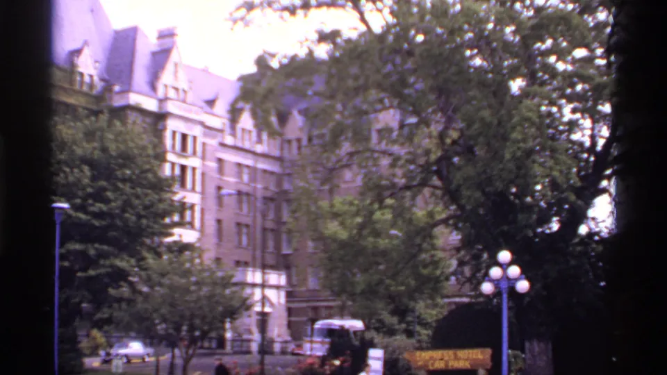 1974 Canada frame of The Empress hotel exterior seen across trees and street furniture, with cars and a parking sign visible.