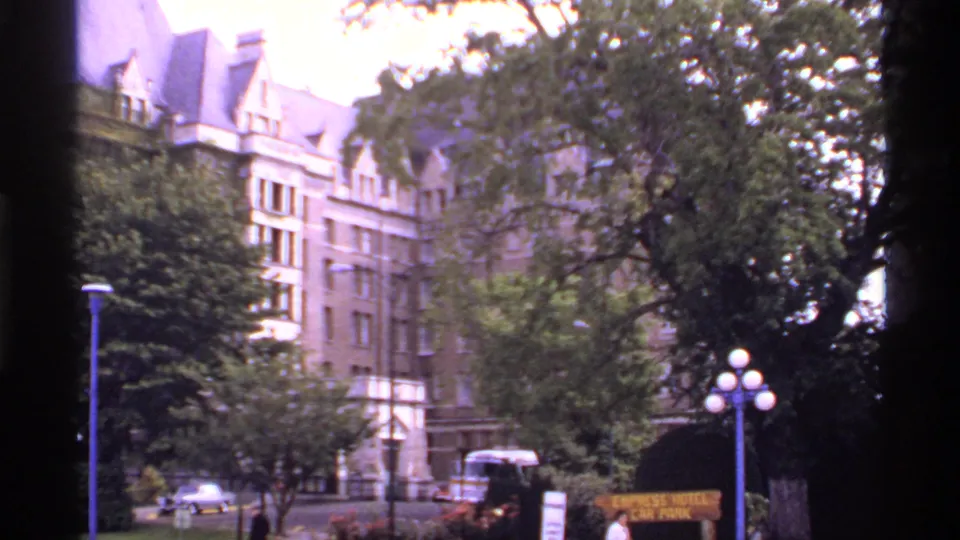1974 Canada 8mm view of the hotel exterior through trees, with vehicles, globe street lamps, and a parking sign at street level.
