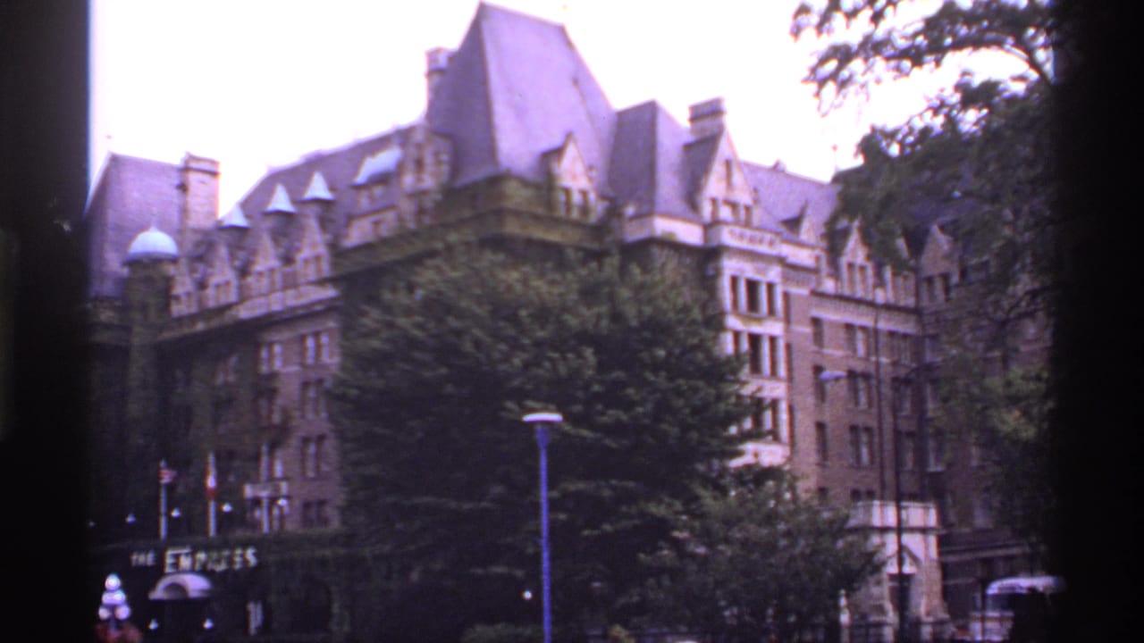 Historic brown building with steep roof behind trees and street lamps.