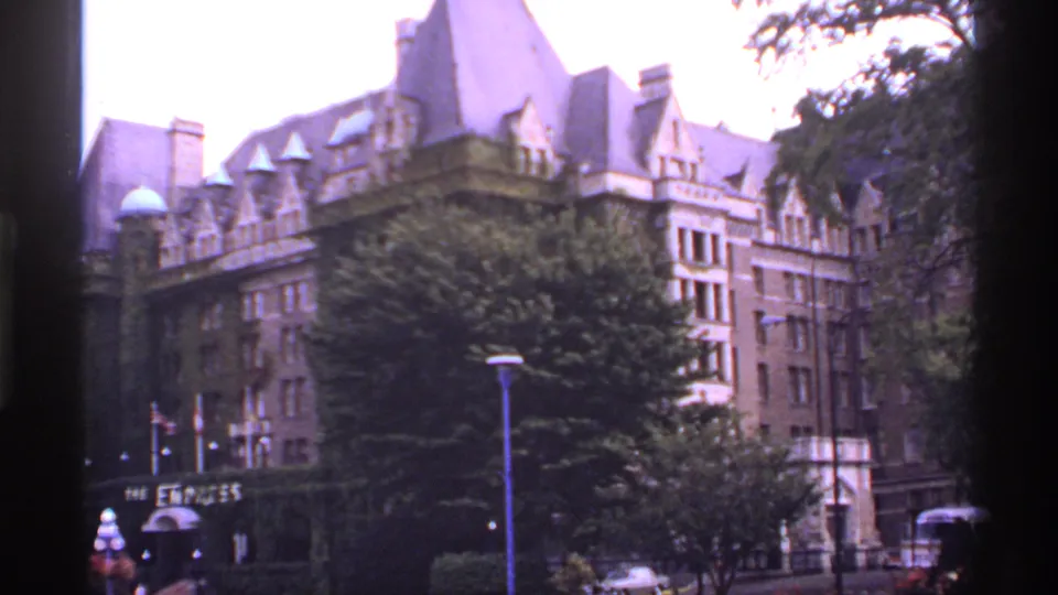 1974 Canadian home-movie frame of a large ornate building, trees in front, flags below, and a car passing near the curb.