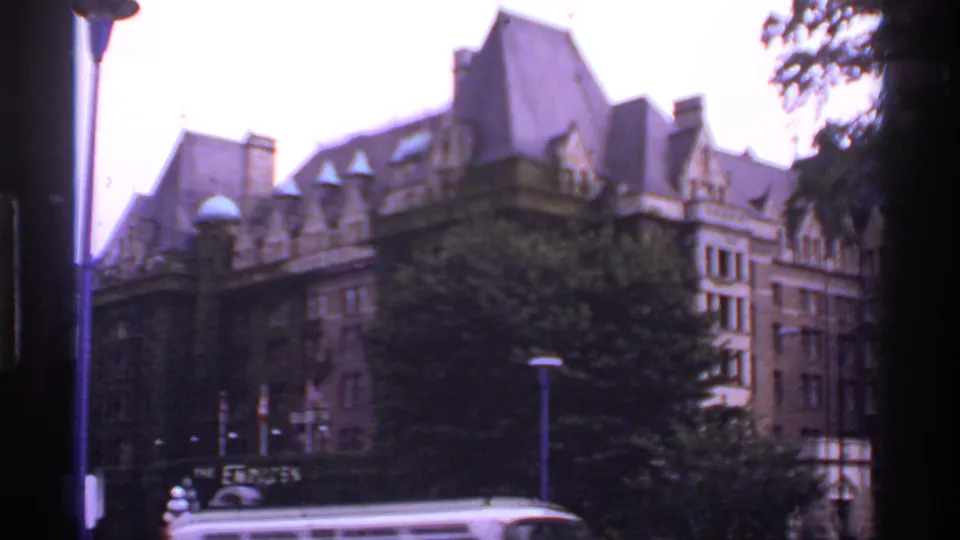 1974 Canadian street view of the brown building and trees, with a light-colored vehicle passing through the lower foreground.