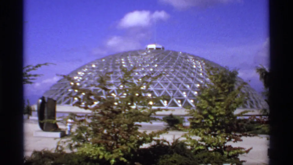 1974 Canadian 8mm frame showing a metallic geodesic conservatory dome under blue sky, partly screened by trees and shrubs.