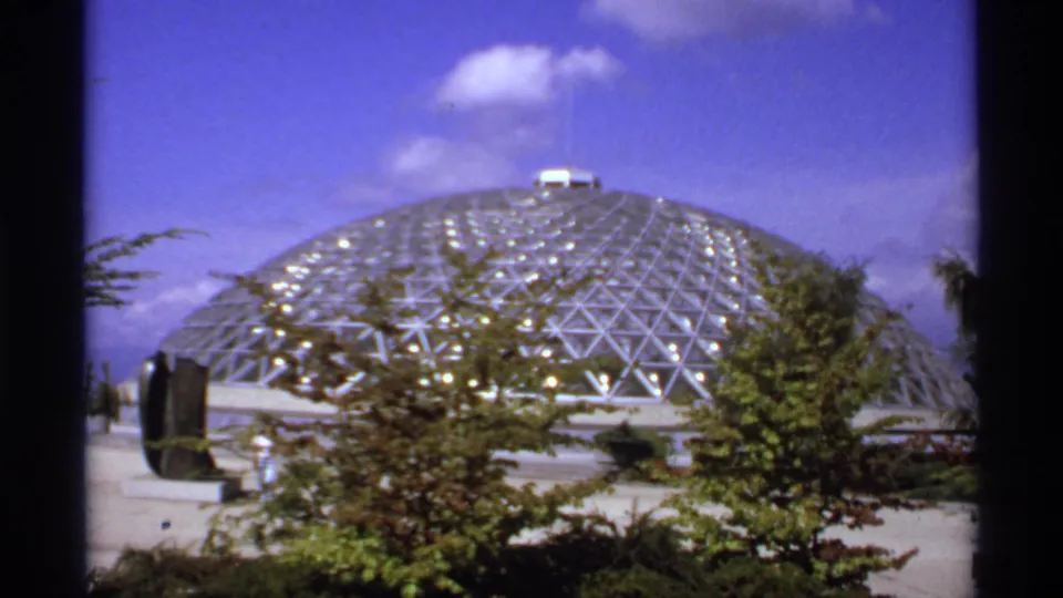 1974 Canadian 8mm frame of a glass-and-metal conservatory dome seen across landscaping, with blue sky and a small cloud above.
