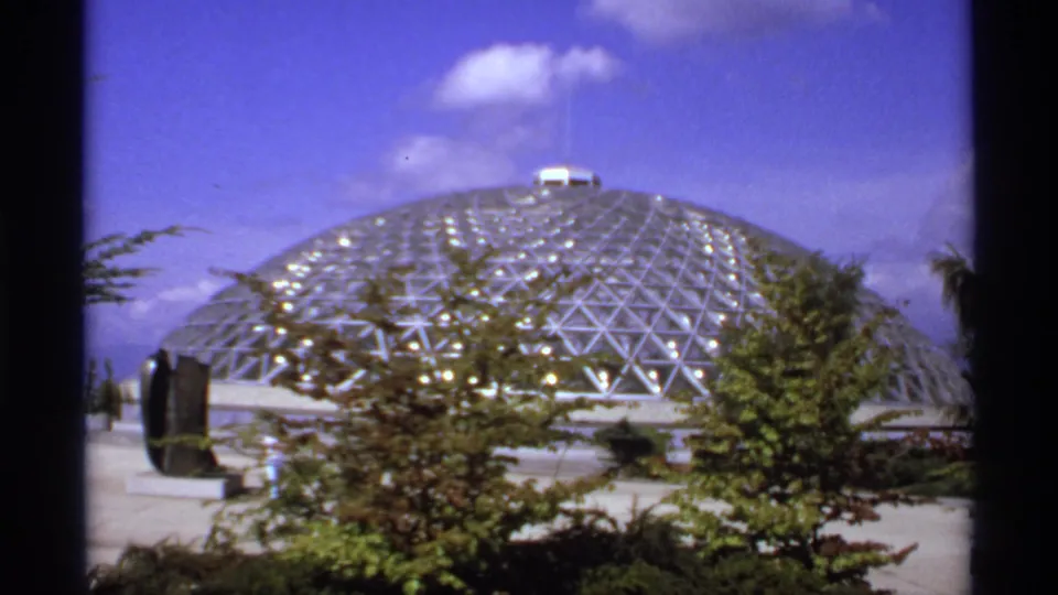 1974 Canadian 8mm frame showing a glass-and-metal geodesic dome beyond trees and a paved outdoor area.