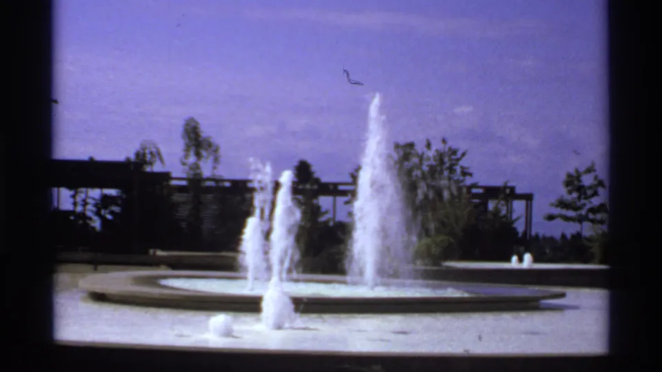 1974 Canadian 8mm frame of a circular fountain spraying water in a landscaped public outdoor setting.