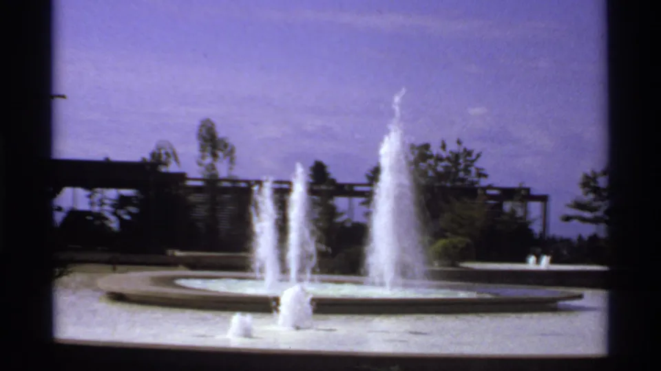 1974 Canadian 8mm frame of a landscaped plaza fountain with tall water sprays and trees beyond.