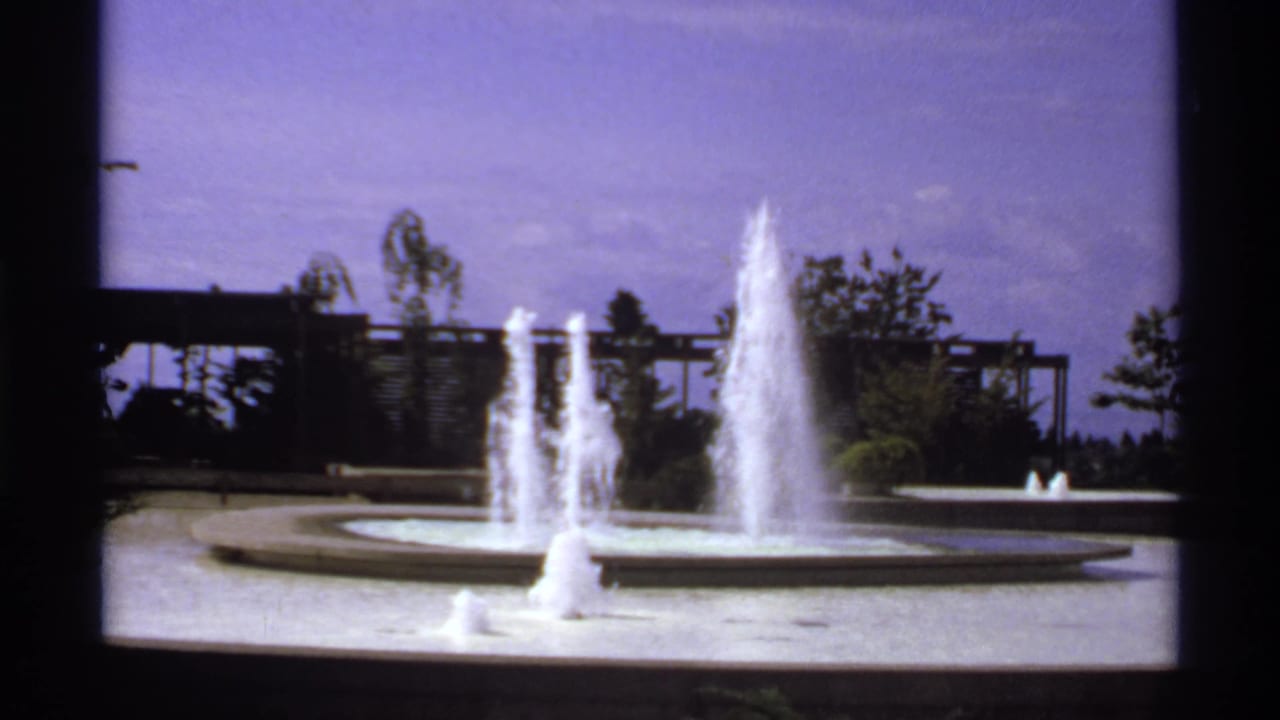 Circular fountain with several water jets, trees, and a trellis-like structure beyond the pool.