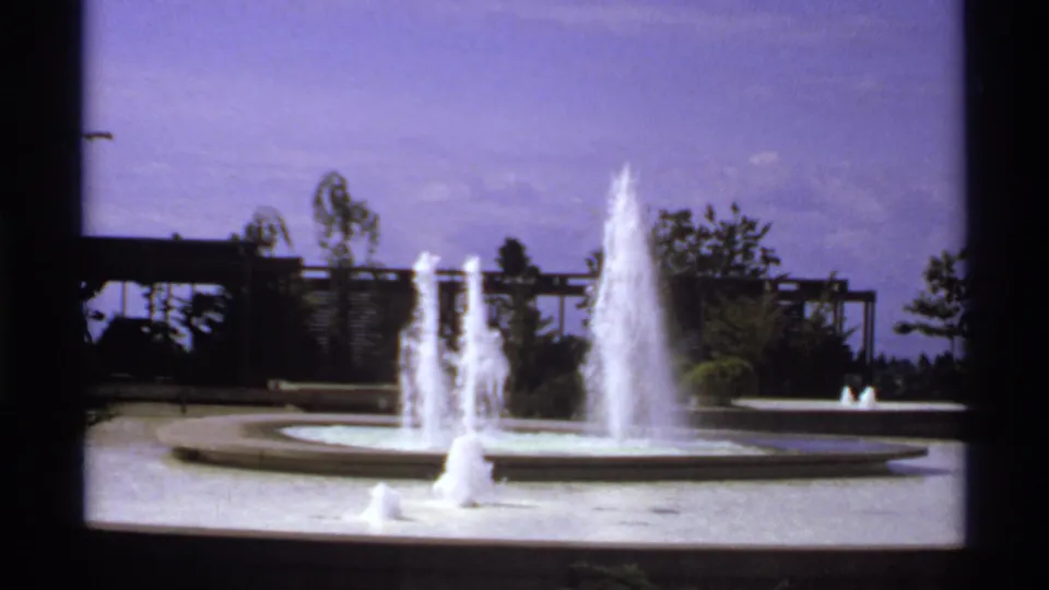 1974 Canada 8mm frame showing a circular fountain with multiple water jets, trees, and a modernist trellis-like background.