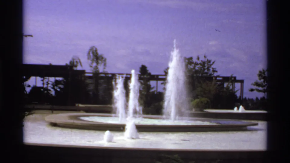 1974 Canada 8mm view of fountain sprays rising from a circular pool, with trees and a trellis-like structure in the background.
