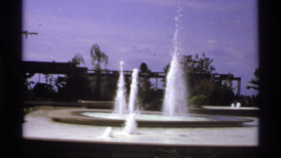 1974 Canada 8mm frame of a circular fountain basin with bright water jets, trees, and a purple-blue sky cast.