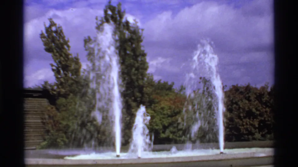 1974 Canadian 8mm footage shows a landscaped park fountain with multiple water jets, autumn trees, shrubs, and a wooden structure at left.