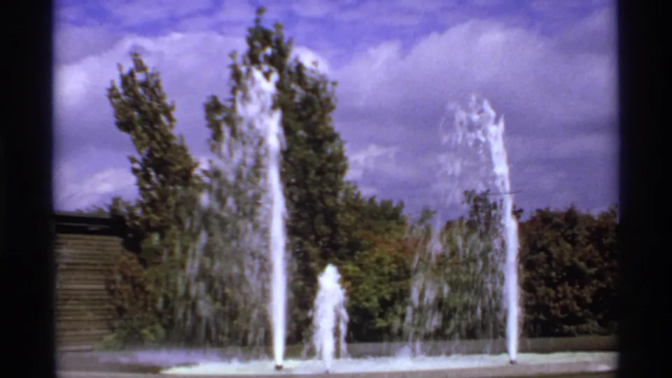 A vintage 8mm park fountain scene in Canada, with tall water jets, surrounding trees, shrubs, and a cloudy purple-blue sky.