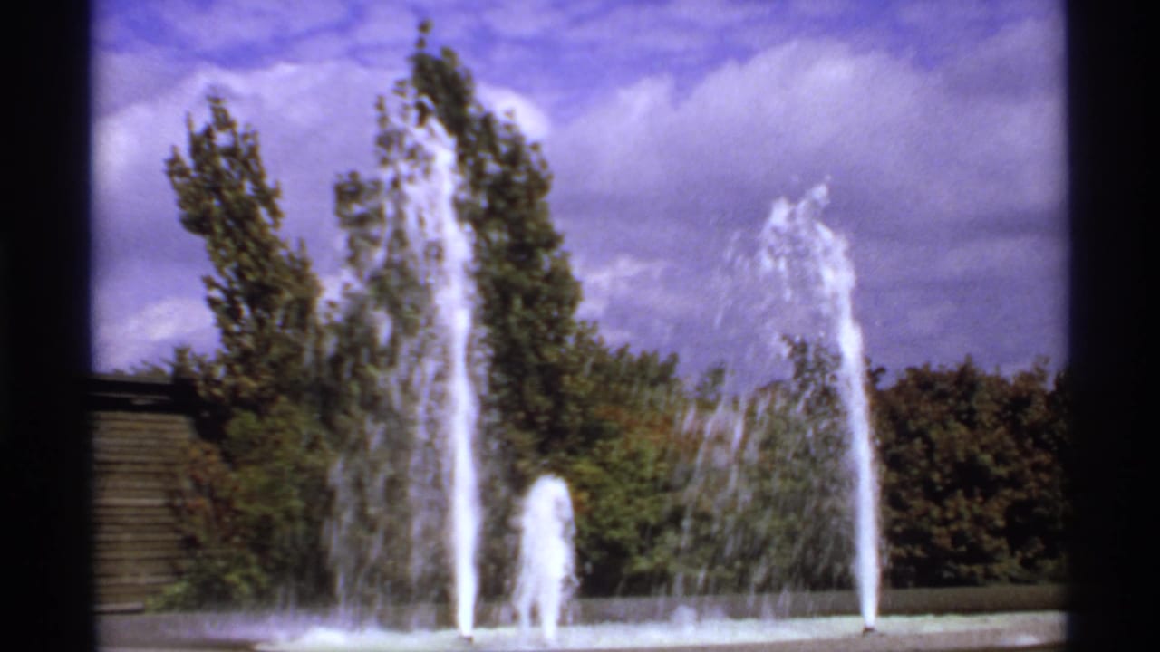 1974 8mm Canada frame showing multiple fountain jets rising before green trees under a partly cloudy sky.