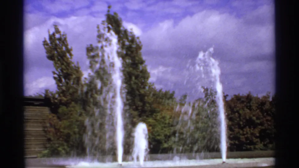 1974 8mm Canada frame showing multiple fountain jets rising before green trees under a partly cloudy sky.