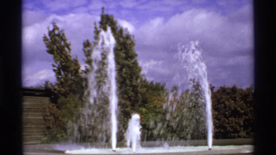 1974 8mm Canada frame of fountain spray patterns against tall trees and a partly cloudy sky.