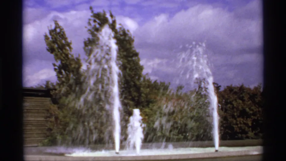 1974 8mm Canada frame with tall water jets, a foamy fountain plume, and dark green trees in the background.