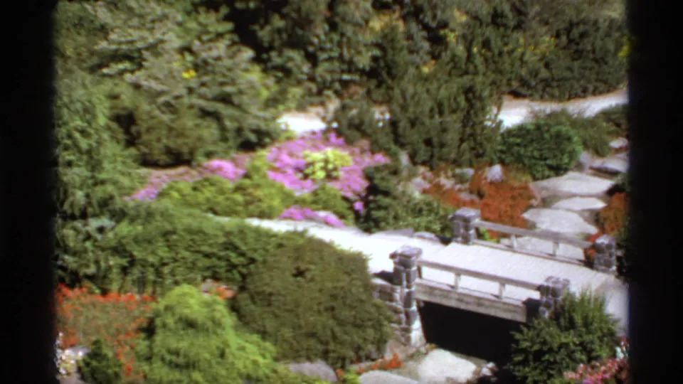 1974 Canadian 8mm frame with a small ornamental bridge, stone paths, shrubs, and bright flower beds.