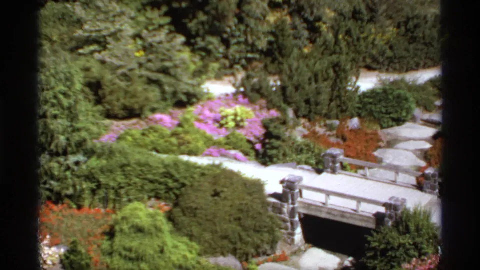 1974 Canadian home-movie frame showing a landscaped garden, low bridge, stone walkway, shrubs, and colorful flowers.