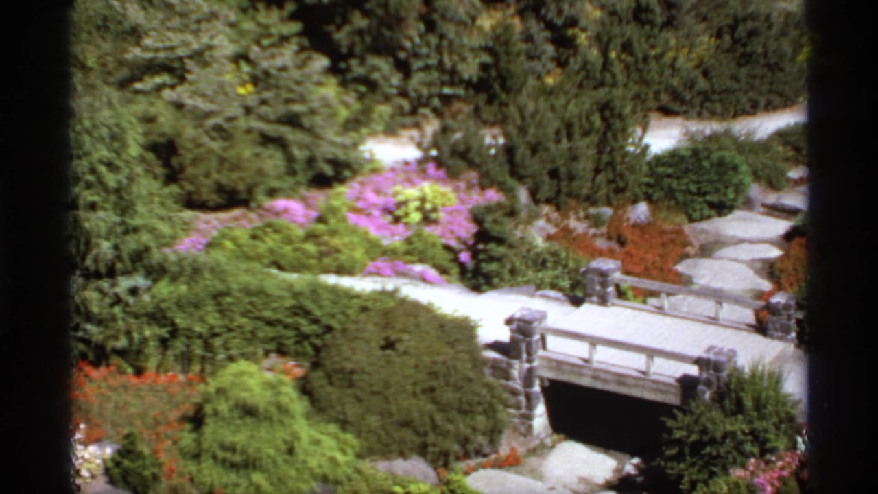 Garden path and stone bridge surrounded by shrubs, trees, and pink flowering plants.