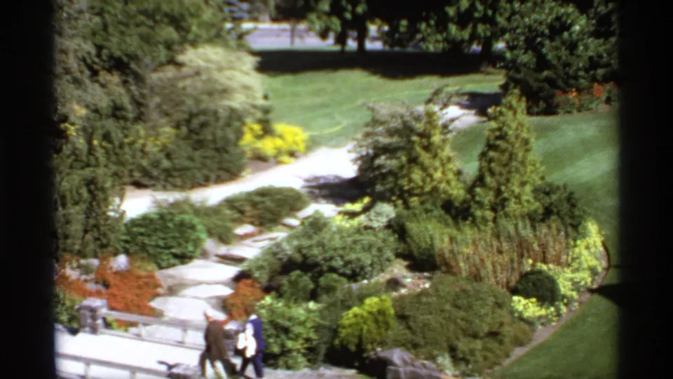 1974 Canadian 8mm frame showing an elderly couple strolling beside a stone bridge in a landscaped garden.