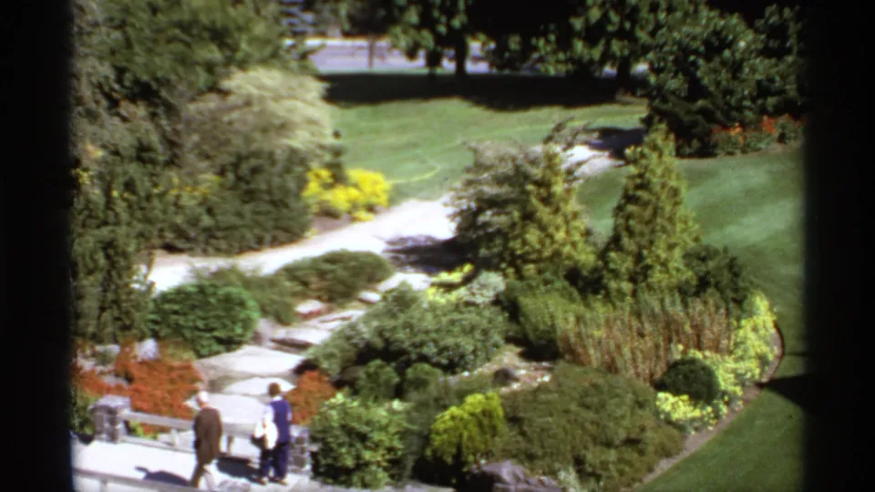1974 Canadian home-movie frame with an elderly couple walking through a green garden of paths, shrubs, rocks, and flowers.