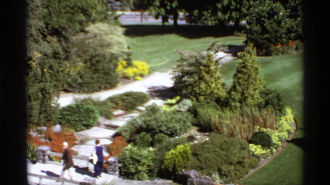Garden paths and green lawns seen through a dark window frame, with two visitors walking below.