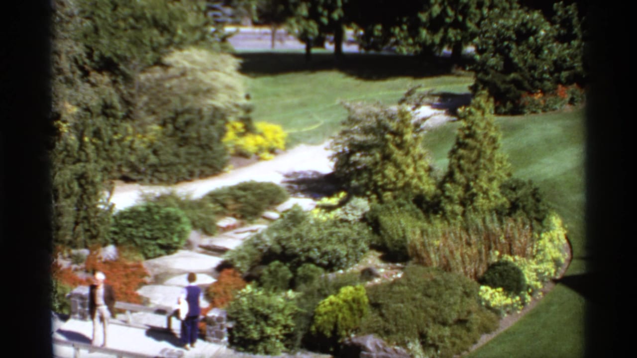 Two visitors walk through a landscaped park with lawns, shrubs, trees, and pale stone paths.