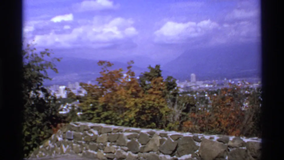 A Canadian scenic overlook in 1974, with city buildings, trees, stonework, and mountains under a purple-tinged sky.