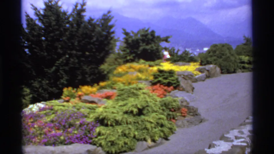 1974 Canadian garden overlook with bright flower beds, manicured shrubs, a curving path, trees, and distant mountains.