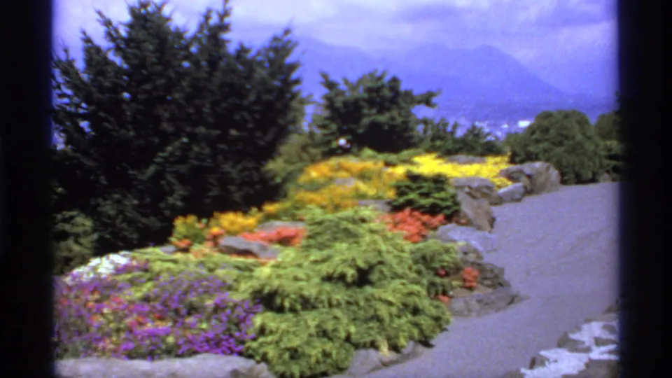 1974 Canada 8mm view of a landscaped garden path with vivid flower beds, evergreen shrubs, rocks, trees, and distant mountains.