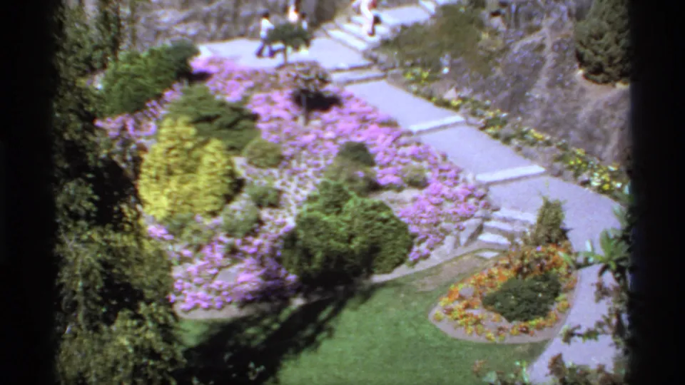 1974 Canada 8mm frame looking down over landscaped paths, purple flower beds, shrubs, lawn, stairs, and distant visitors.