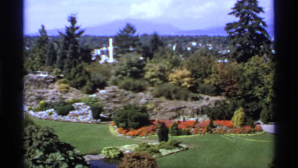1974 Canada 8mm wide view across a landscaped garden, red flower border, trees, distant buildings, and hazy mountains.