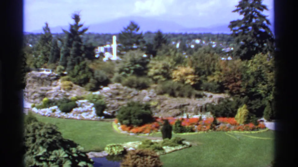 1974 Canada frame with manicured lawn, red flowerbeds, shrubs, tall evergreens, distant buildings, and mountains under bright daylight.