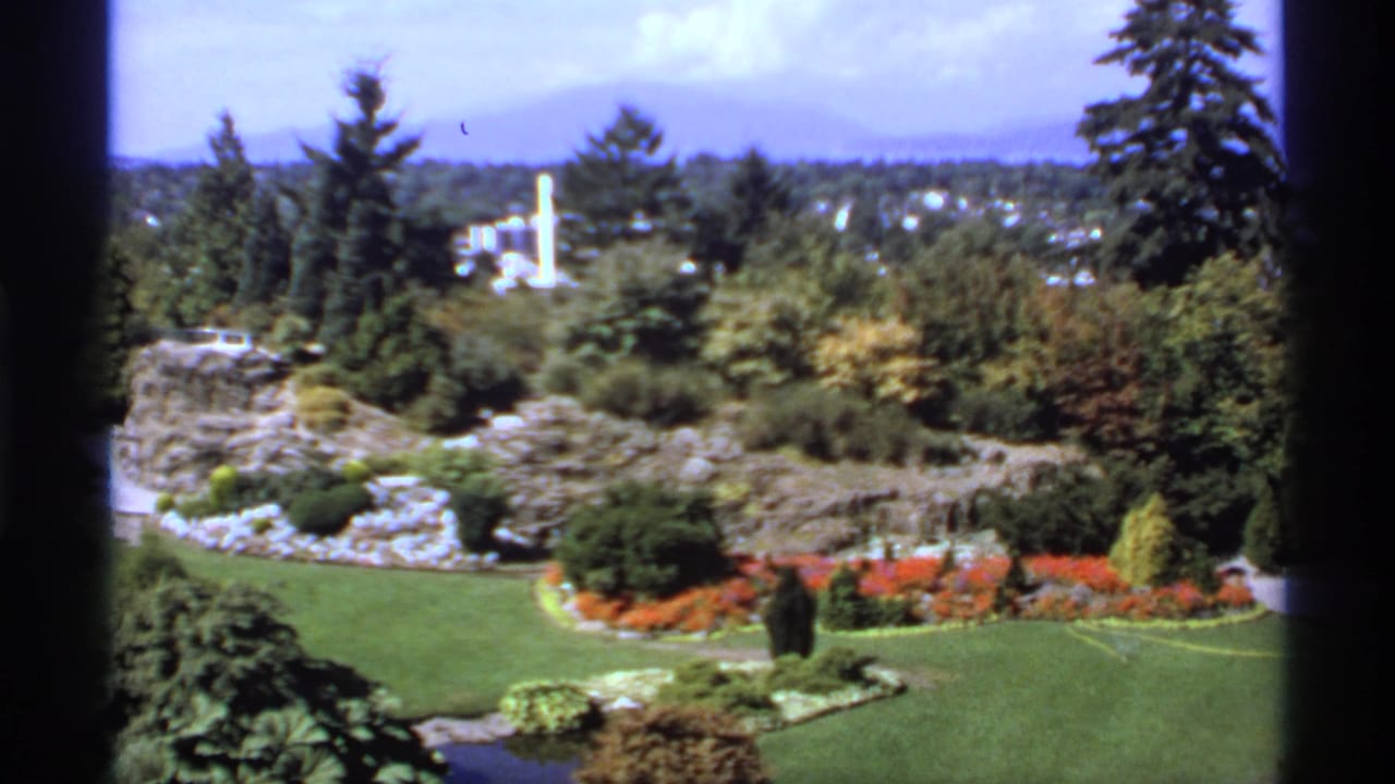 High view over a landscaped garden with lawn, flower beds, trees, water, and distant hills.