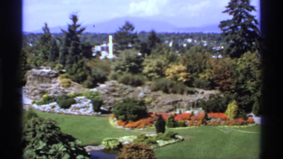 1974 Canadian 8mm frame showing a high-angle view of lawns, red flower beds, trees, a water feature, and a distant white monument.