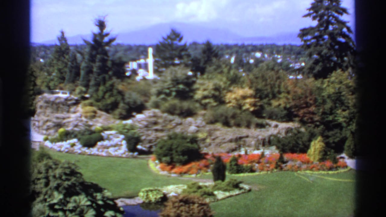 High garden view with clipped grass, flower beds, shrubs, trees, water, and a white structure in the distance.