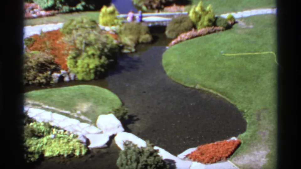 1974 Canadian 8mm frame looking down on a garden water feature, stone paths, flower beds, a small bridge, and tiny visitors in the distance.