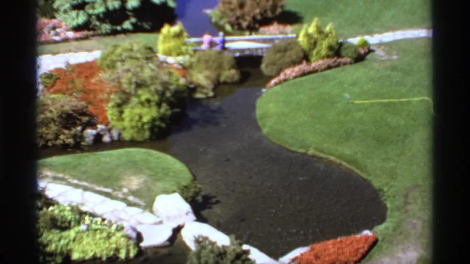 1974 Canadian garden frame with a winding dark water feature, bright lawns, planting beds, stone paths, a bridge, and small visitors far away.