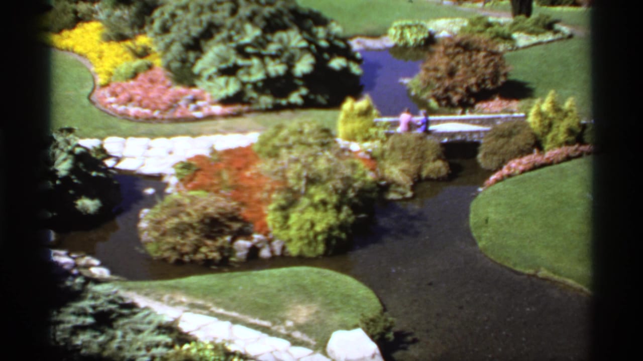 Two distant people stand on a small bridge above a brook in a landscaped garden.