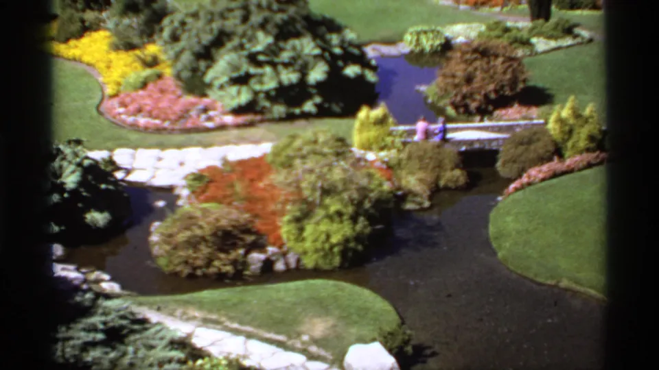 1974 Canadian 8mm frame looking across a landscaped brook toward two people on a bridge, with bright flowerbeds and trimmed lawns nearby.