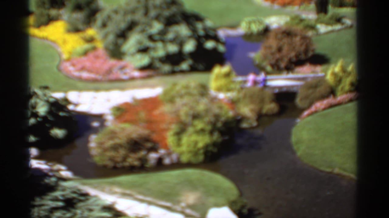 1974 Canada 8mm view of landscaped garden grounds with curving lawn, dark pond water, shrubs, flowerbeds, and a small bridge.