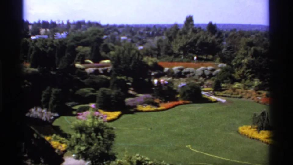 1974 Canada 8mm elevated view over landscaped garden grounds with a large lawn, flowerbeds, dark trees, rocks, paths, and distant treeline.
