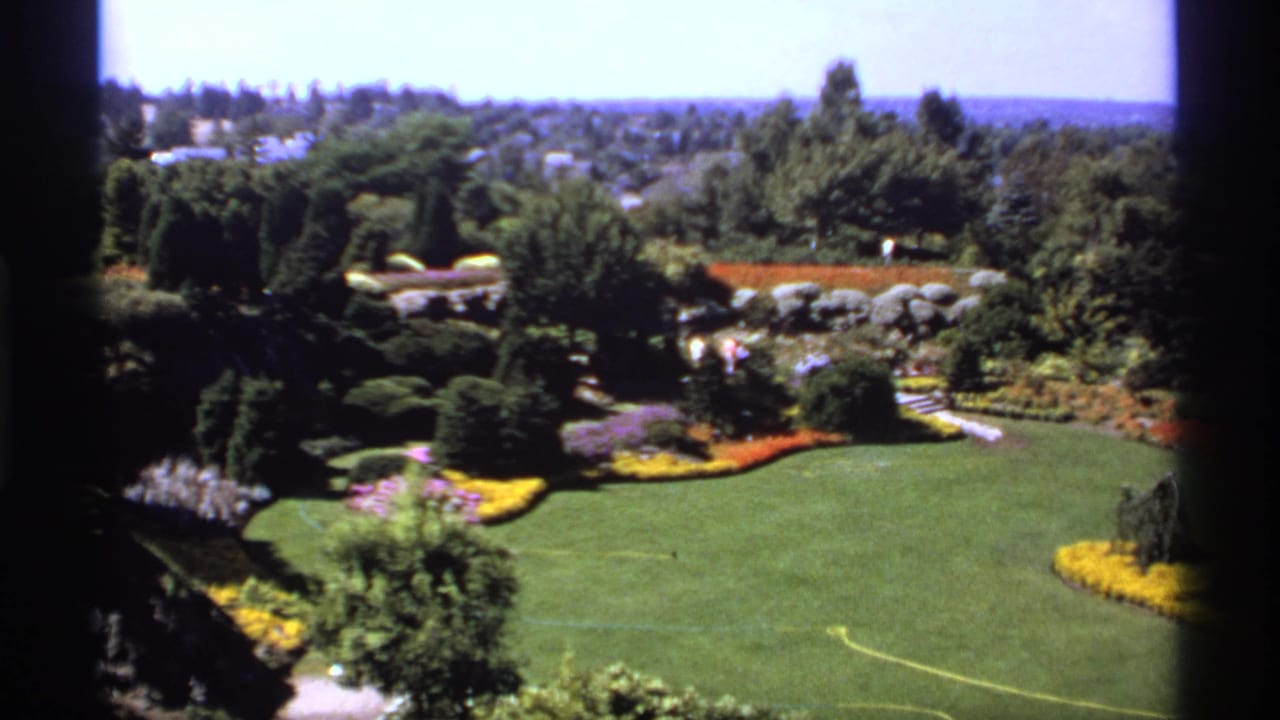 Elevated view over a landscaped garden with lawn, flowerbeds, hedges, trees, and distant houses.