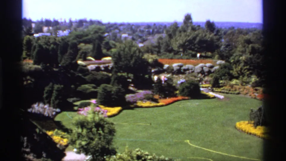 1974 Canadian 8mm frame showing a sunlit landscaped park with a broad lawn, colorful beds, clipped shrubs, and distant suburban rooftops.