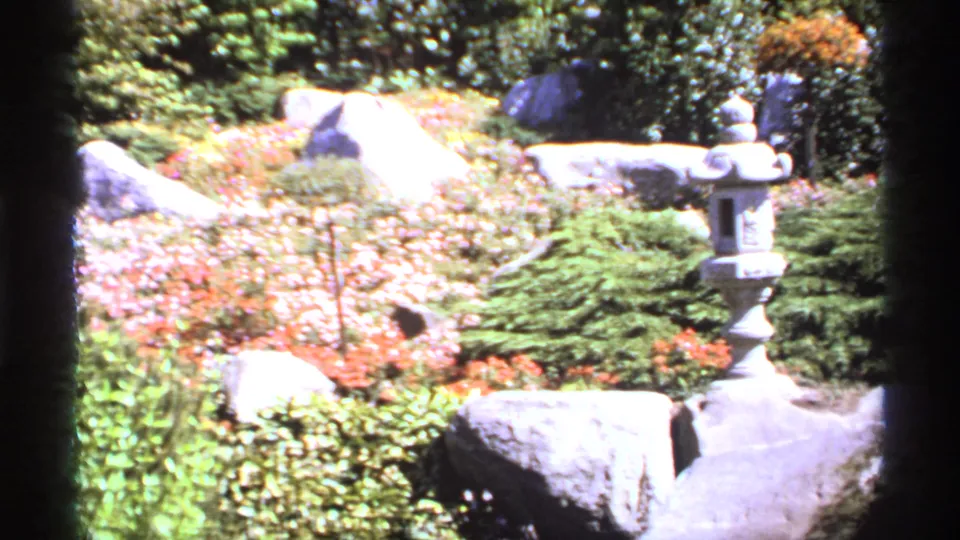 1974 Canadian 8mm frame showing a Japanese-style garden area with a stone lantern, large rocks, low shrubs, flowers, and a paved path.